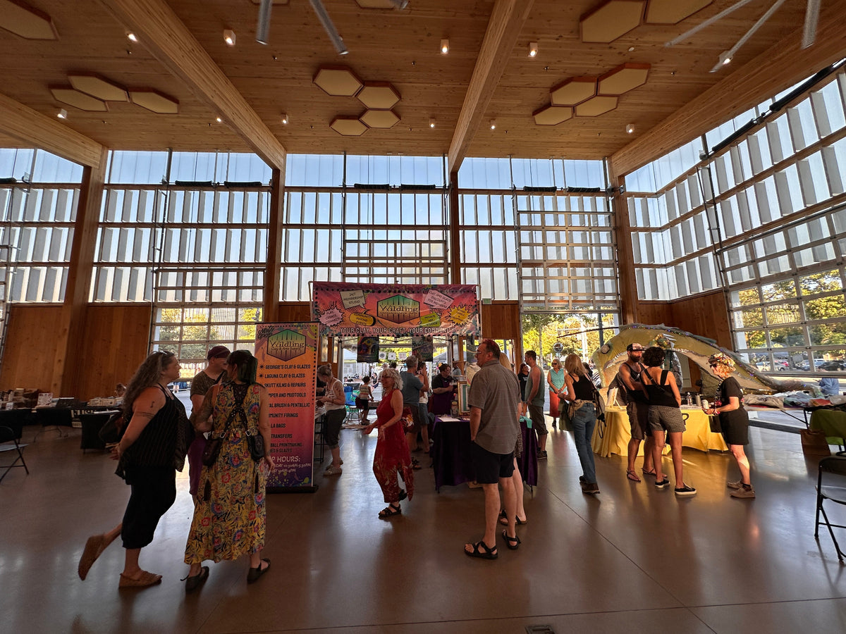 A bustling indoor event or fair taking place in a large modern hall with high windows, wood ceiling panels, and several people walking around and standing at booths.