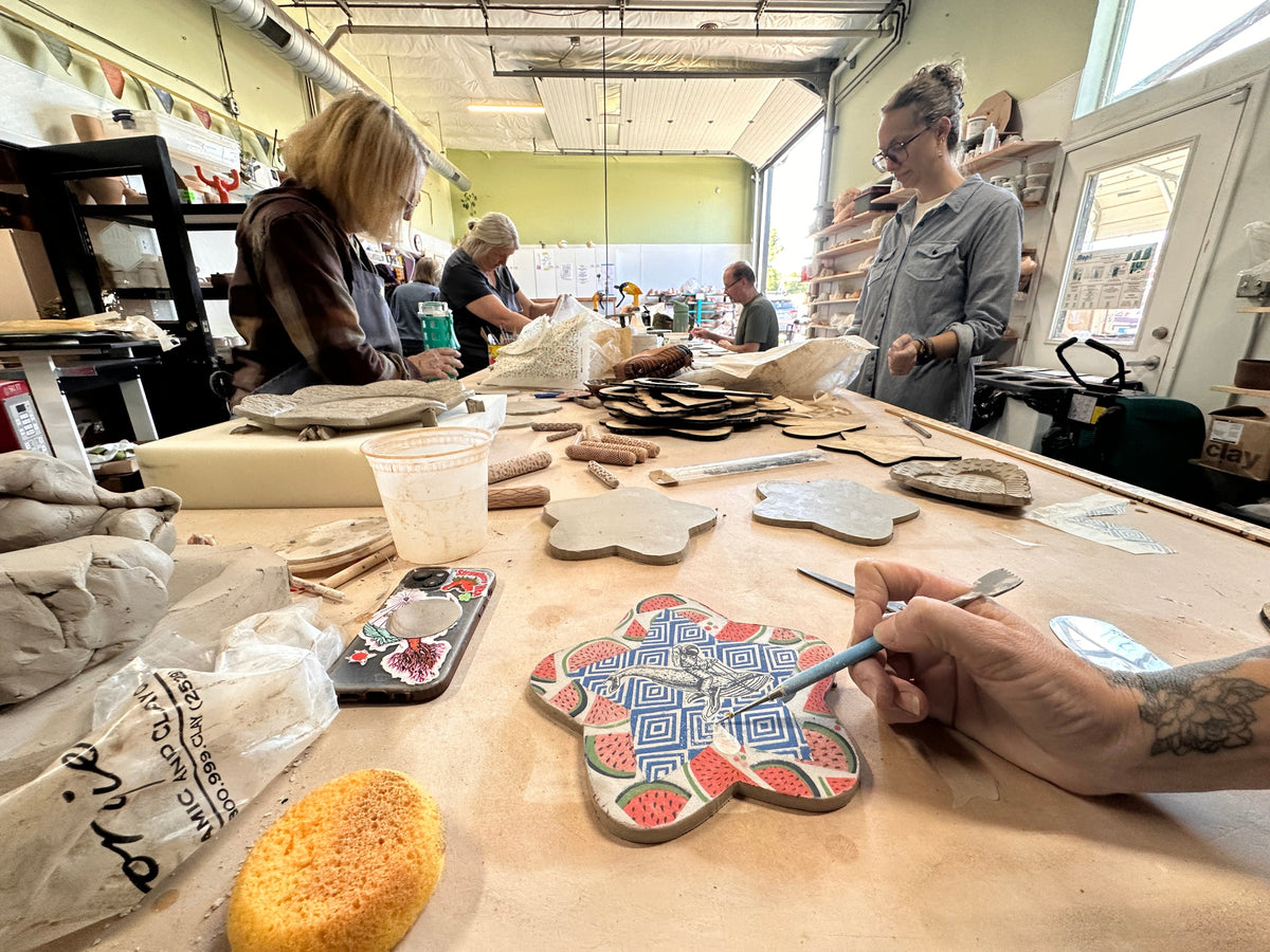 A person's hand applies underglaze transfer to a handmade clay tile with red, blue, and white geometric patterns in a pottery studio. Other artisans and clay pieces are visible in the background workspace.