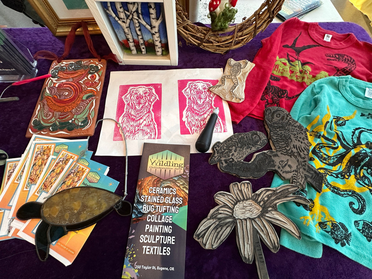 A tabletop display of various handmade crafts, textiles, and art supplies, including a package of Wildpine Ceramic Stained Glass Bug Tufting, pink dog block prints, colorful t-shirts, and wooden bird and flower cutouts on a purple fabric background.