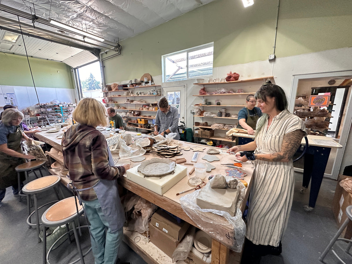 A pottery studio with several people working around a large clay table, shaping and glazing ceramic pieces in a bright indoor workshop.