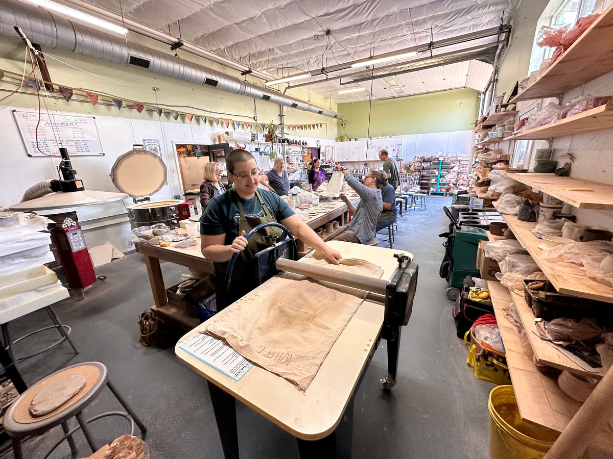 A person working at a pottery table in a studio, shaping a beige clay slab with a wooden tool. The workshop contains shelves, pottery, and other artisans working in the background.