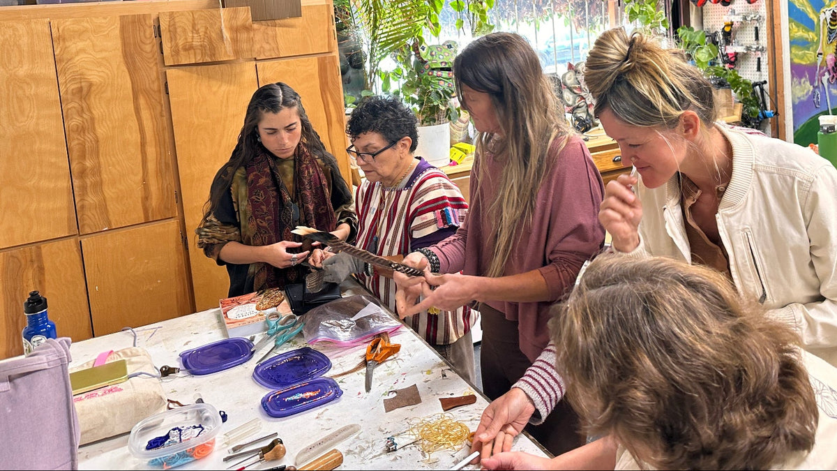 A group of people gathered around a table in a creative studio, working on a craft project with beads, tools, and materials. The room is decorated with artwork, plants, and shelves, creating a vibrant and artistic atmosphere.