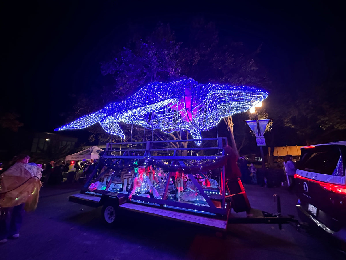 A nighttime parade scene with a trailer decorated with a large whale-shaped wire light display glowing in blue and white, surrounded by festival lights and people on a dark street.