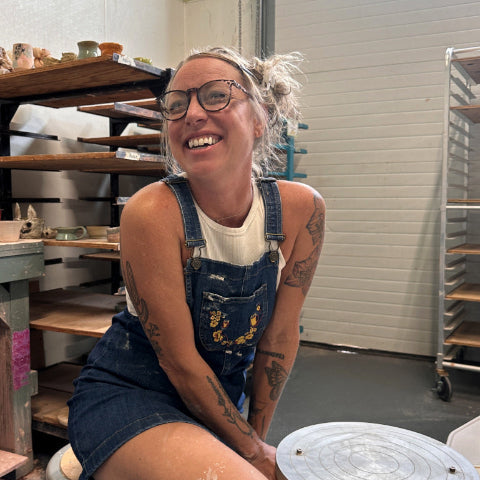 Allie wearing glasses and tattoos sits on a stool in a pottery studio, wearing blue denim overalls over a white t-shirt. A pottery wheel is in the foreground, with shelves and racks in the background.