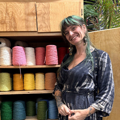 Jayme Vineyard, Co-Founder, with green hair stands smiling in front of an open wooden cabinet filled with colorful spools of thread or yarn. She is wearing a dark tie-dye dress with blue and white patterns.