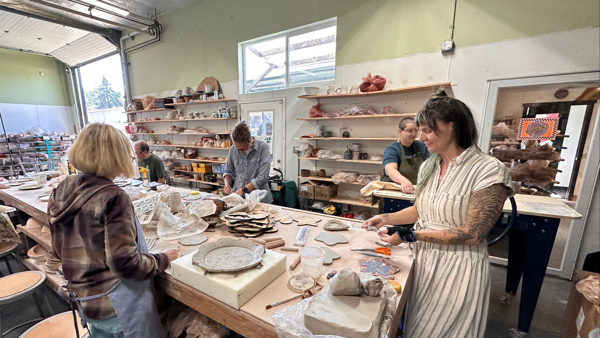A pottery studio with several people working around a large clay table, shaping and glazing ceramic pieces in a bright indoor workshop.