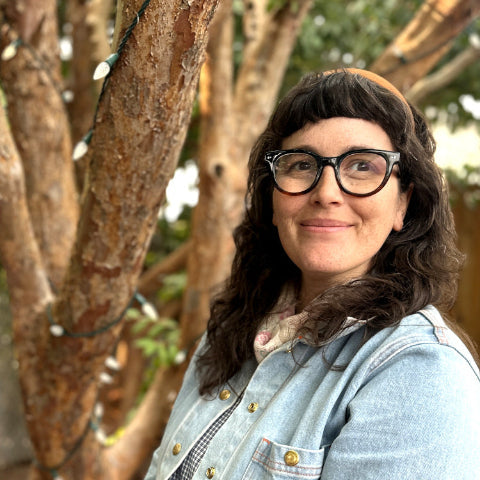 Kate standing with dark hair and glasses wearing a light blue denim jacket over a checkered shirt, standing outdoors in front of tree trunks with string lights.