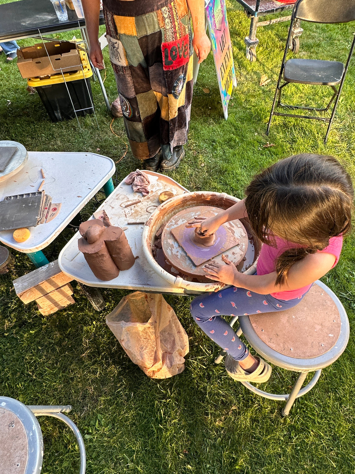 A young girl sits at a pottery wheel, shaping clay outdoors on a grassy lawn. An adult stands nearby, and pottery wheels, tools, and a small table are visible in the scene.