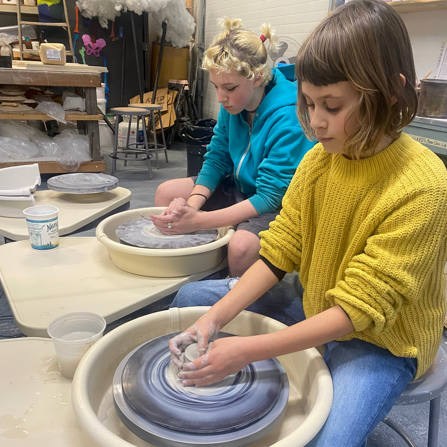 A young girl in a yellow knit sweater sits at a pottery wheel shaping clay, with another person working at a similar wheel in the background inside a pottery studio.