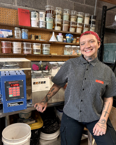 Kyle smiling inside the glaze mixing  workshop behind a countertop, wearing a black-and-white gingham shirt and dark pants. Shelves behind them are filled with many labeled jars and containers, and a blue研 mill machine is visible on the left.