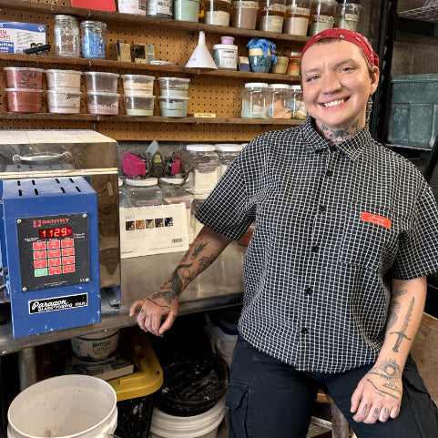 Kyle smiling inside the glaze mixing  workshop behind a countertop, wearing a black-and-white gingham shirt and dark pants. Shelves behind them are filled with many labeled jars and containers, and a blue研 mill machine is visible on the left.