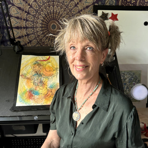 Michelle smiling with short blonde hair sits indoors wearing a dark green short-sleeve button-up shirt, layered necklaces, and bracelets. She is seated in front of artwork and a black equipment case in a room setting.