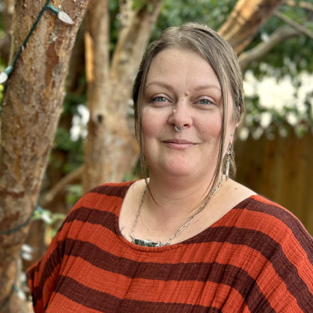Co-Founder, Sarah Root, standing with tattoos wearing a rust-orange and dark brown horizontally striped dress, standing outdoors in front of a wooden fence and trees.